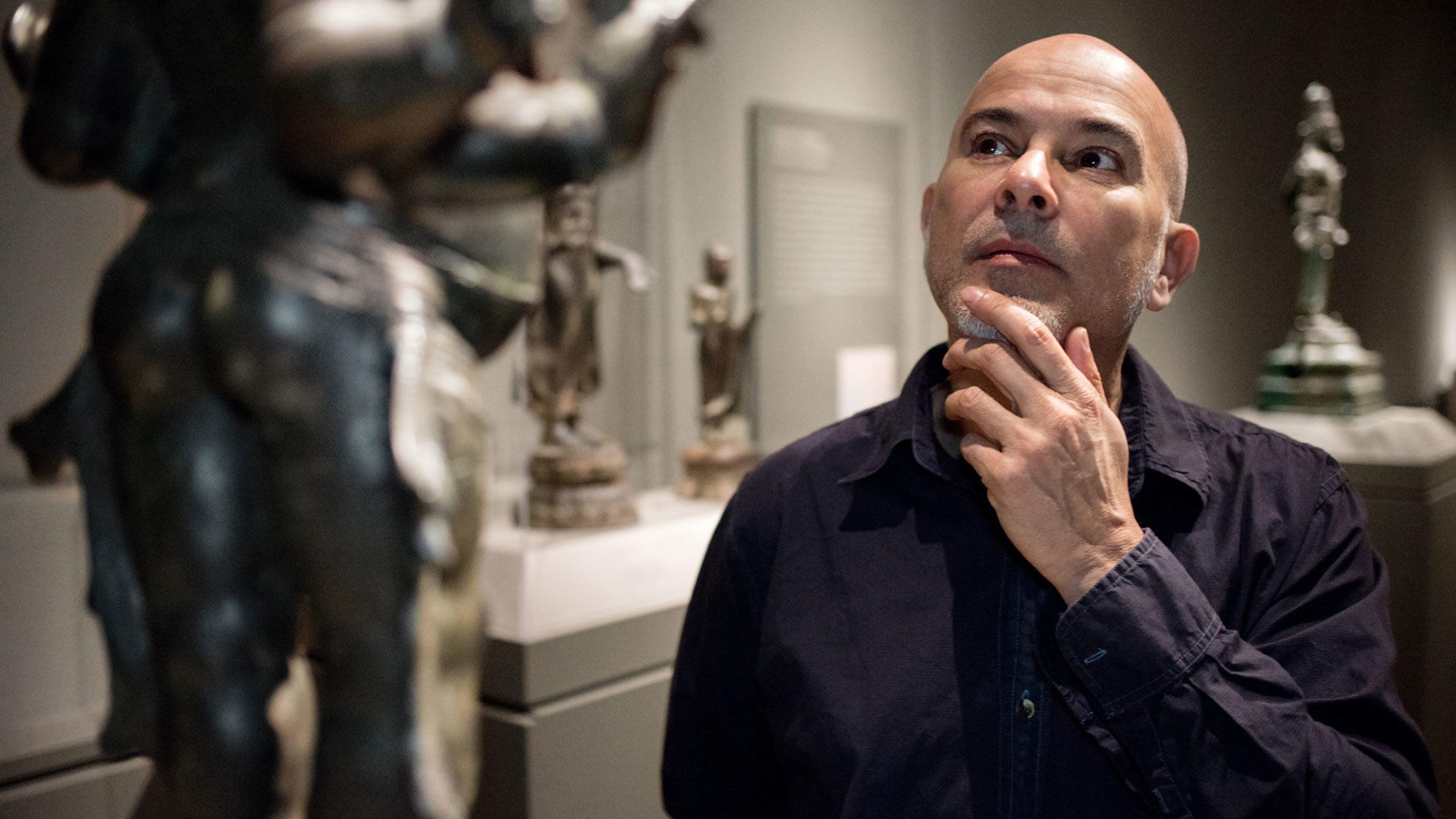 A bald man in a button down shirt looks at a bronze sculpture, holding his hand to his chin.