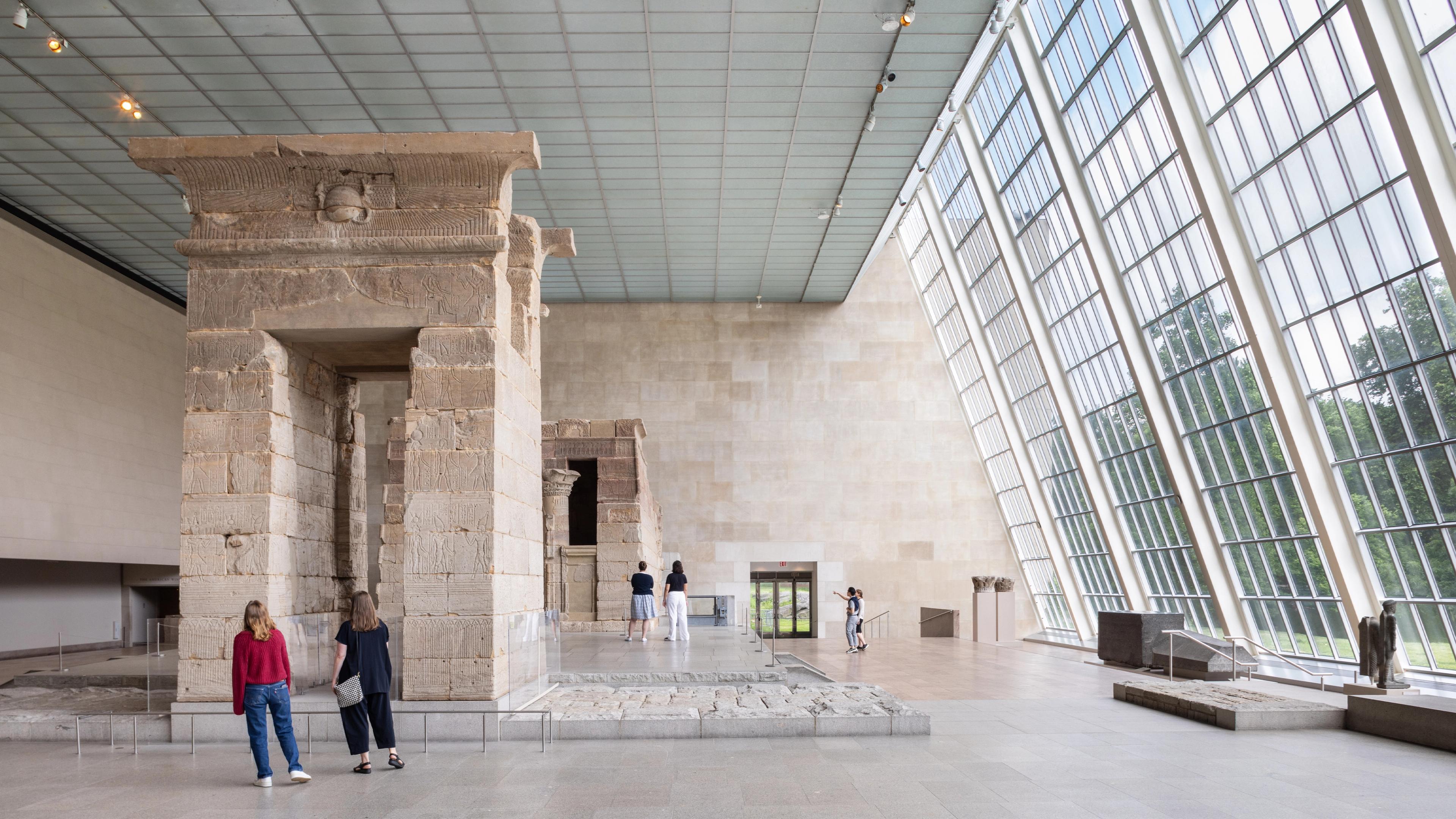 Visitors admire ancient stone structures in a spacious museum hall with large glass window on the right wall that illuminating the space.