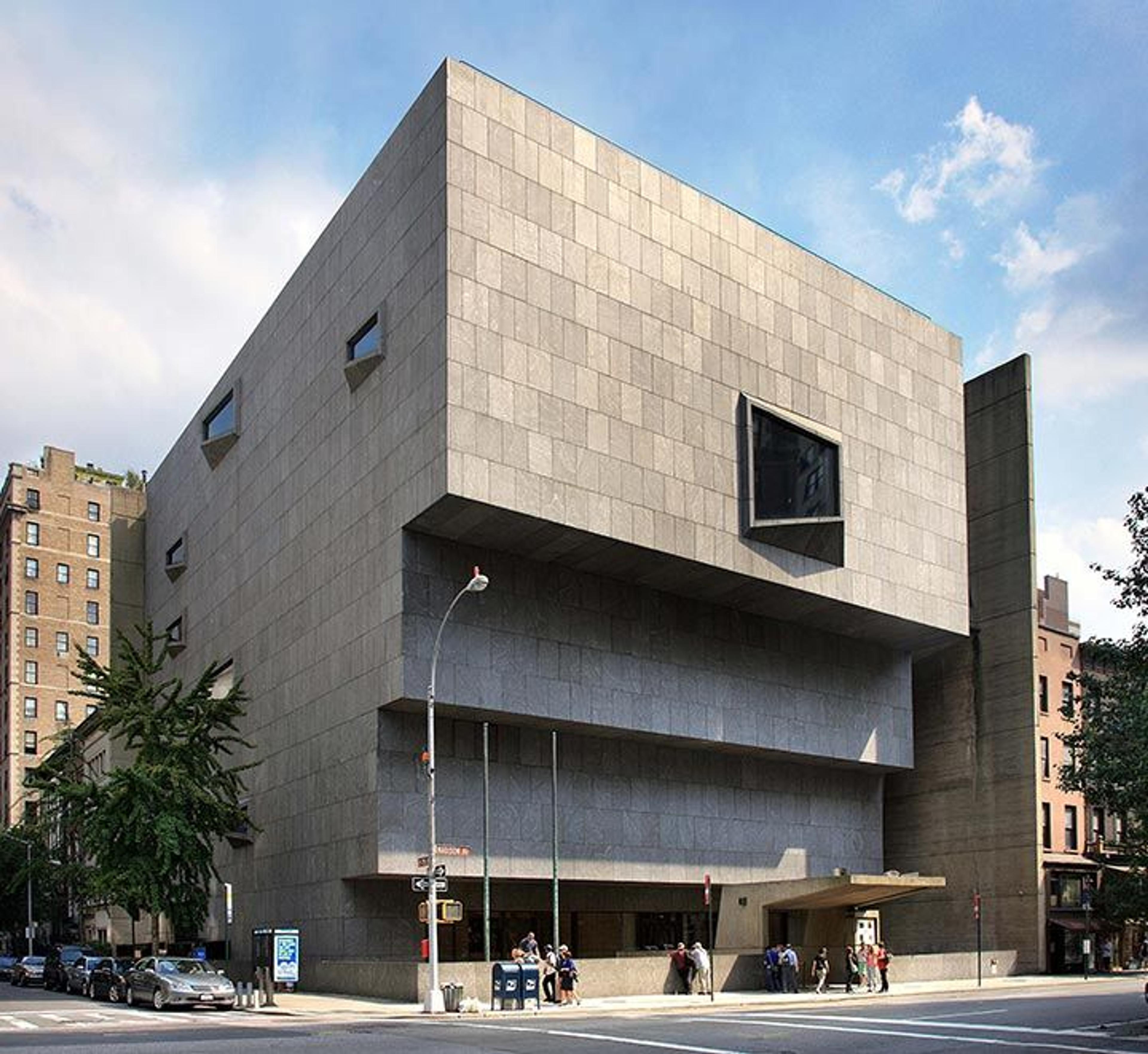 Exterior of a modern, angular concrete building with one large windows toward the top of the front wall, set against a clear sky and urban surroundings.